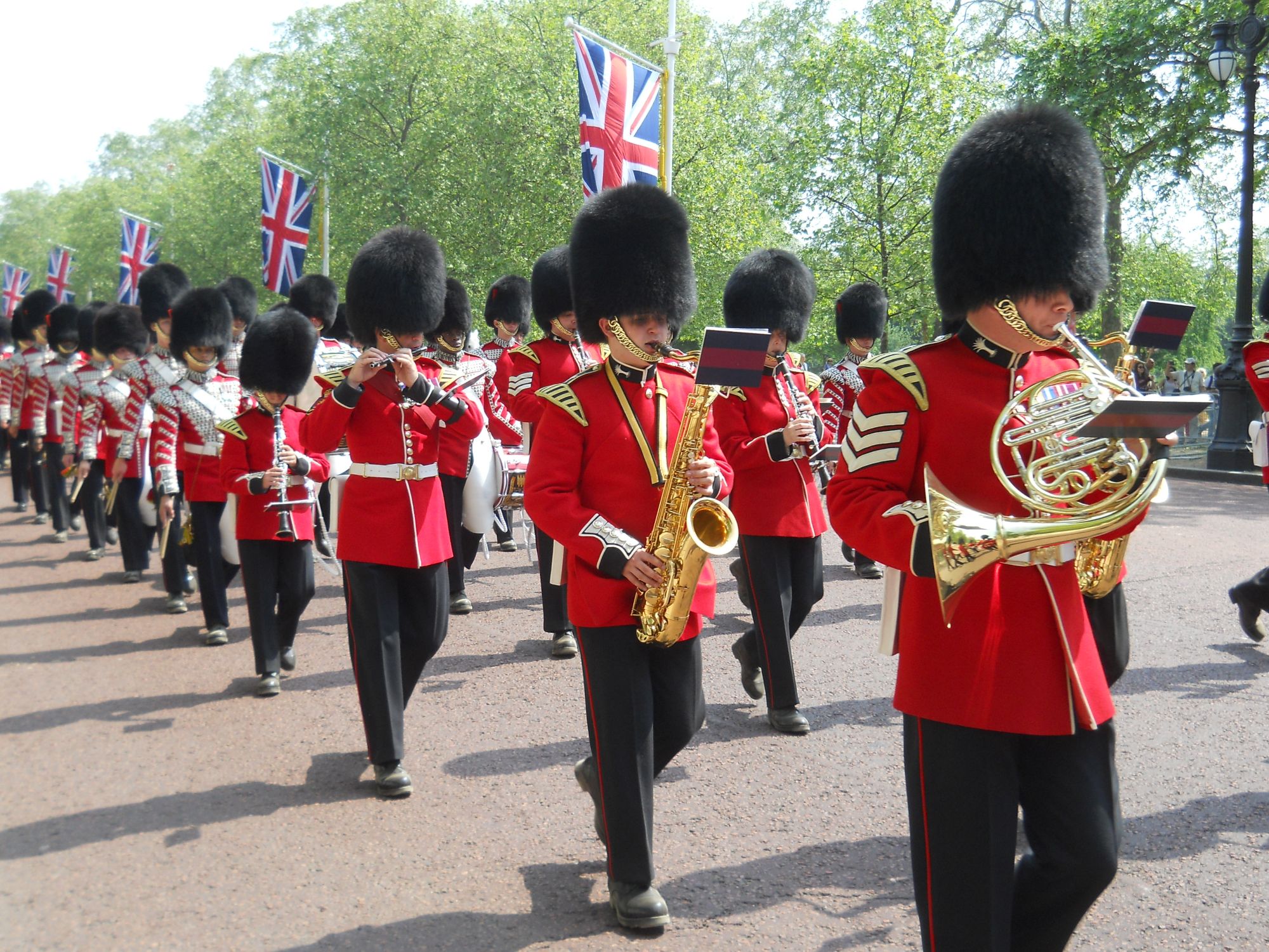 Photo of a military band marching  along The Mall. The soliders were scarlet jackets and black busbys. They are playing a selection of brass and woodwind instruments.